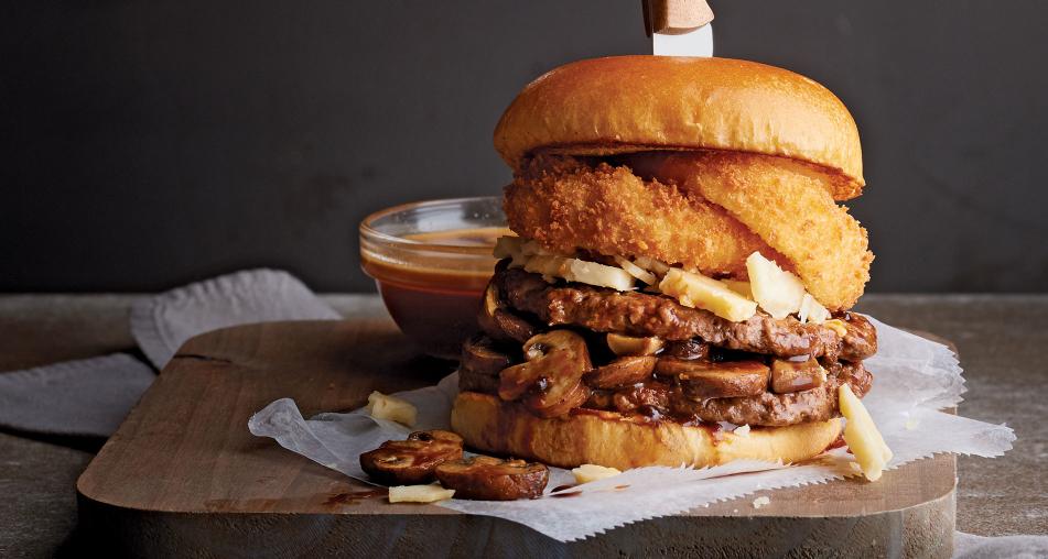 A burger stacked with onion rings, mushrooms, and cheese next to a small bowl of sauce.