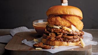 A burger stacked with onion rings, mushrooms, and cheese next to a small bowl of sauce.